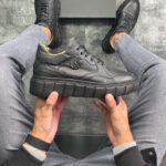 Kožne muške poluduboke cipele u crnoj boji sa visokim crnim đonom. Prikaz bočne strane iz ruke modela u odevnoj kombinaciji.